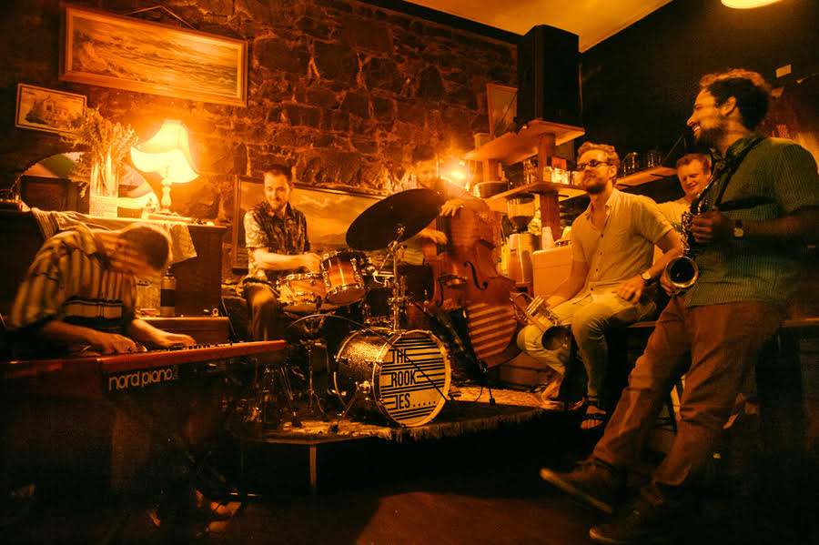 A group of rock musicians plays on a small stage inside a golden-lit bar.