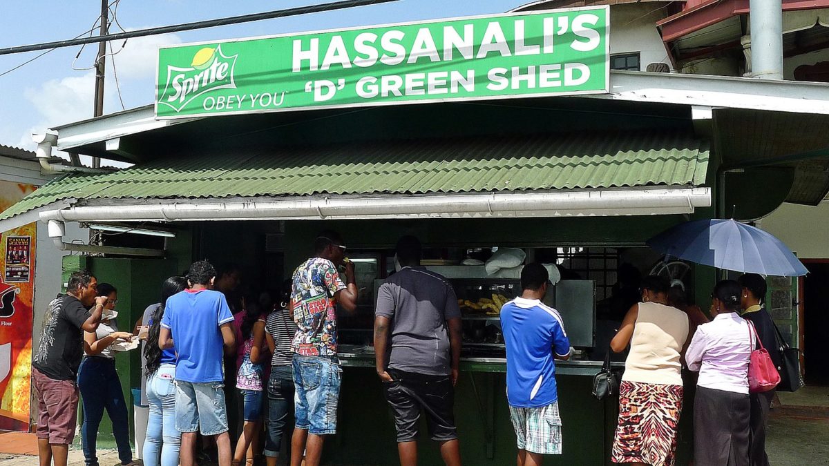 People gathered in front of a green food stand with a large sign reading "Hassanali's"