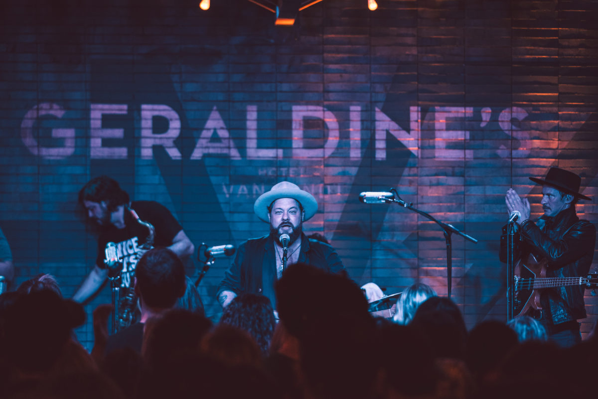 A singer illuminated in blue performs onstage for a crowd in front of a brick wall painted with the phrase "Geraldine's"