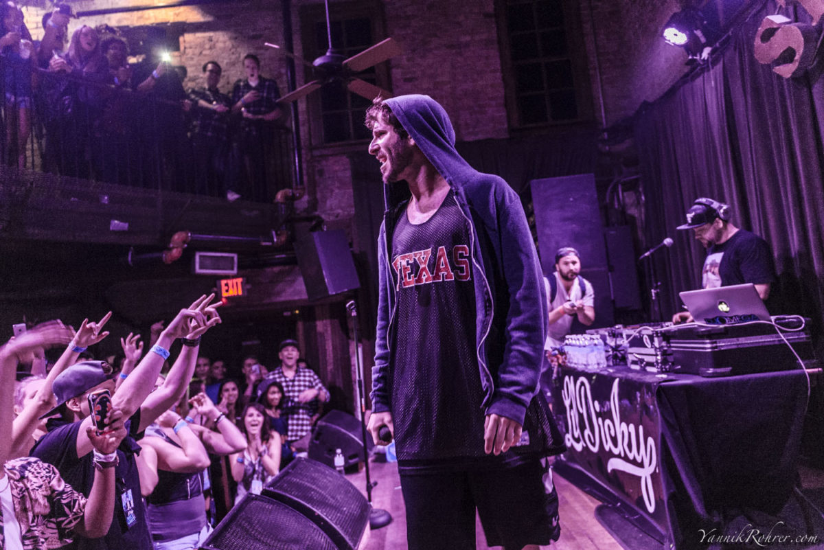 A man in a hoodie standing on a stage illuminated by purple light looks out into a crowd