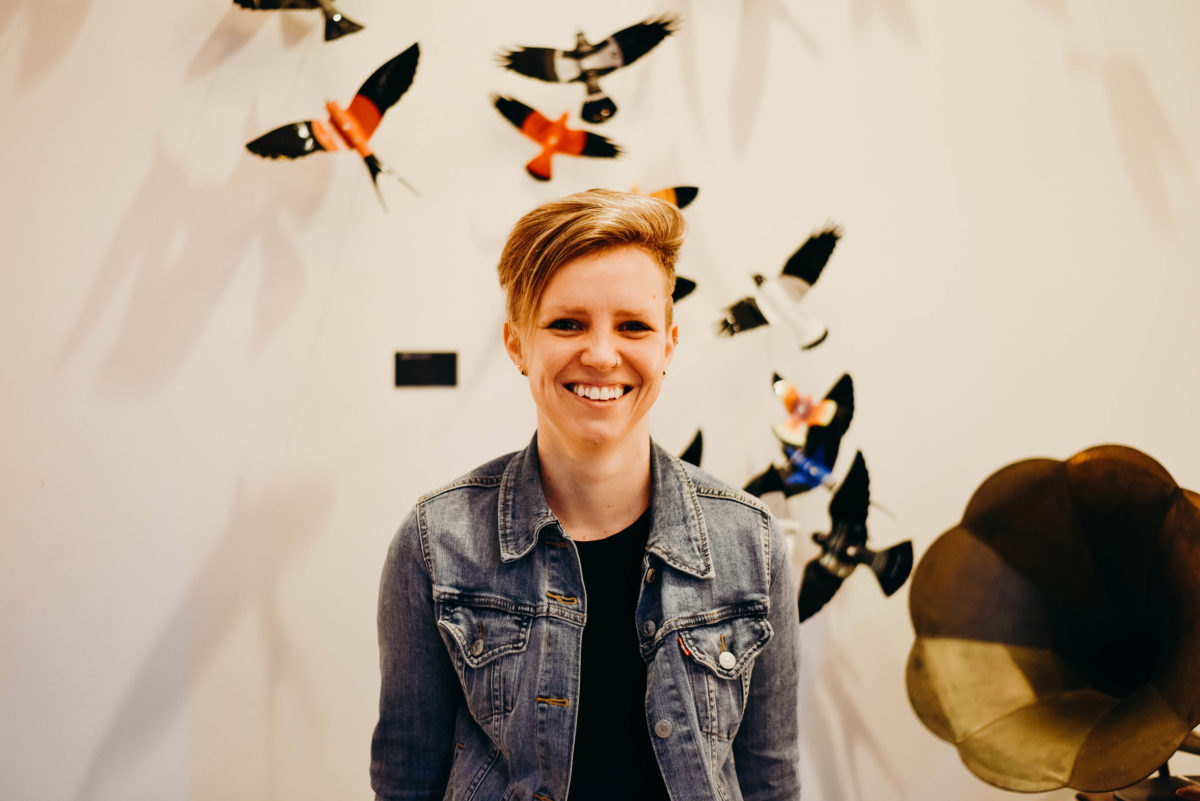 A woman with short blonde hair in a black shirt and denim jacket smiles in front of a white wall decorated with birds