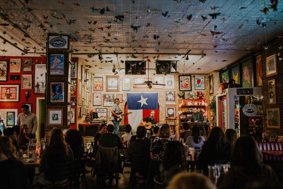 A crowd sits before a stage on which a man plays guitar with the Texas flag hung on the colorful behind him