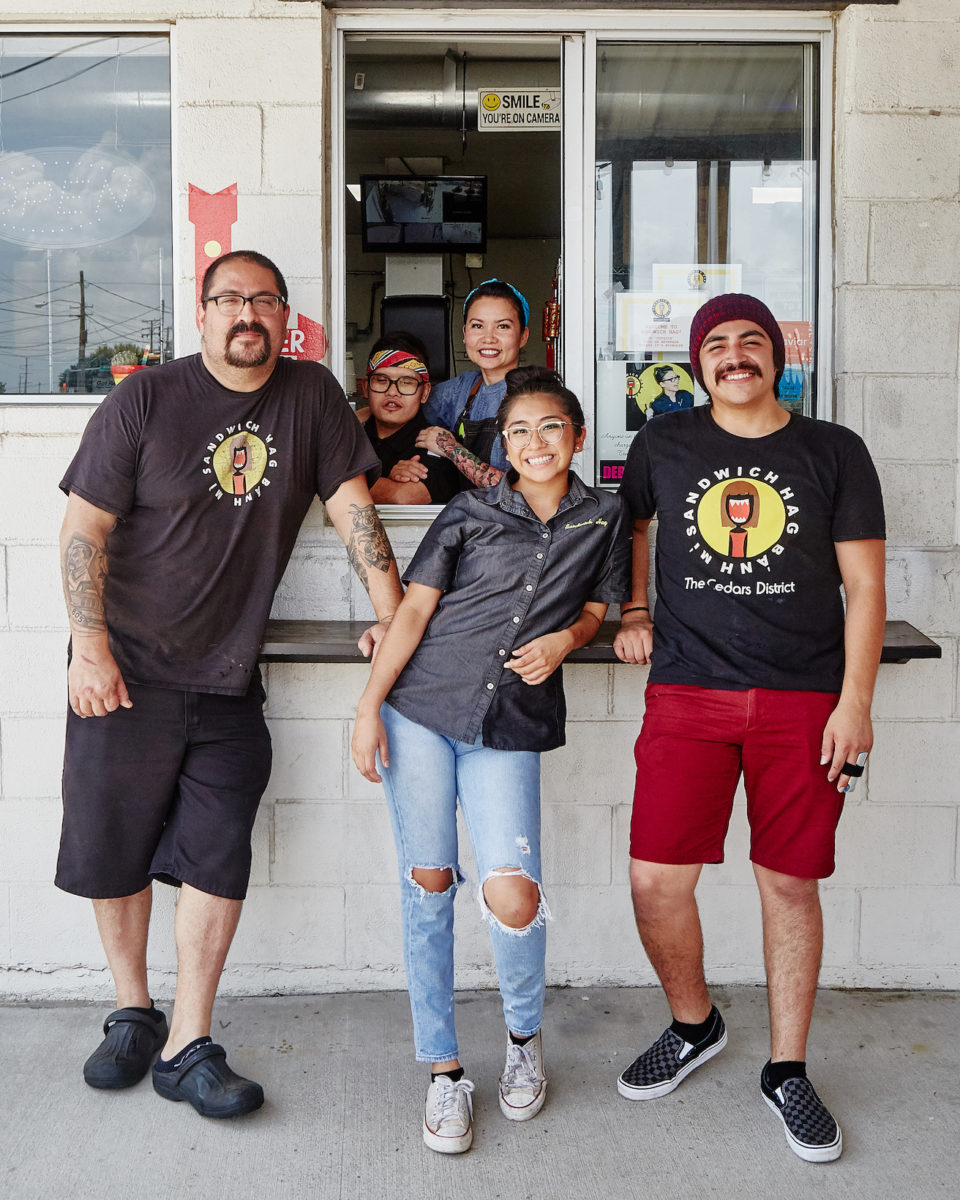 a group of people wearing black shirts from Sandwich Hag stands in front of a white wall with a small window