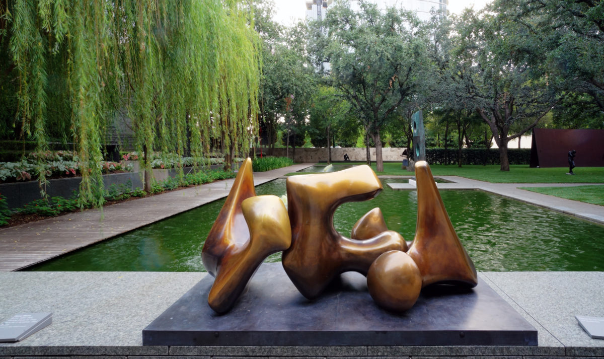 a golden sculpture of multiple shapes with a large green pond and trees in the background