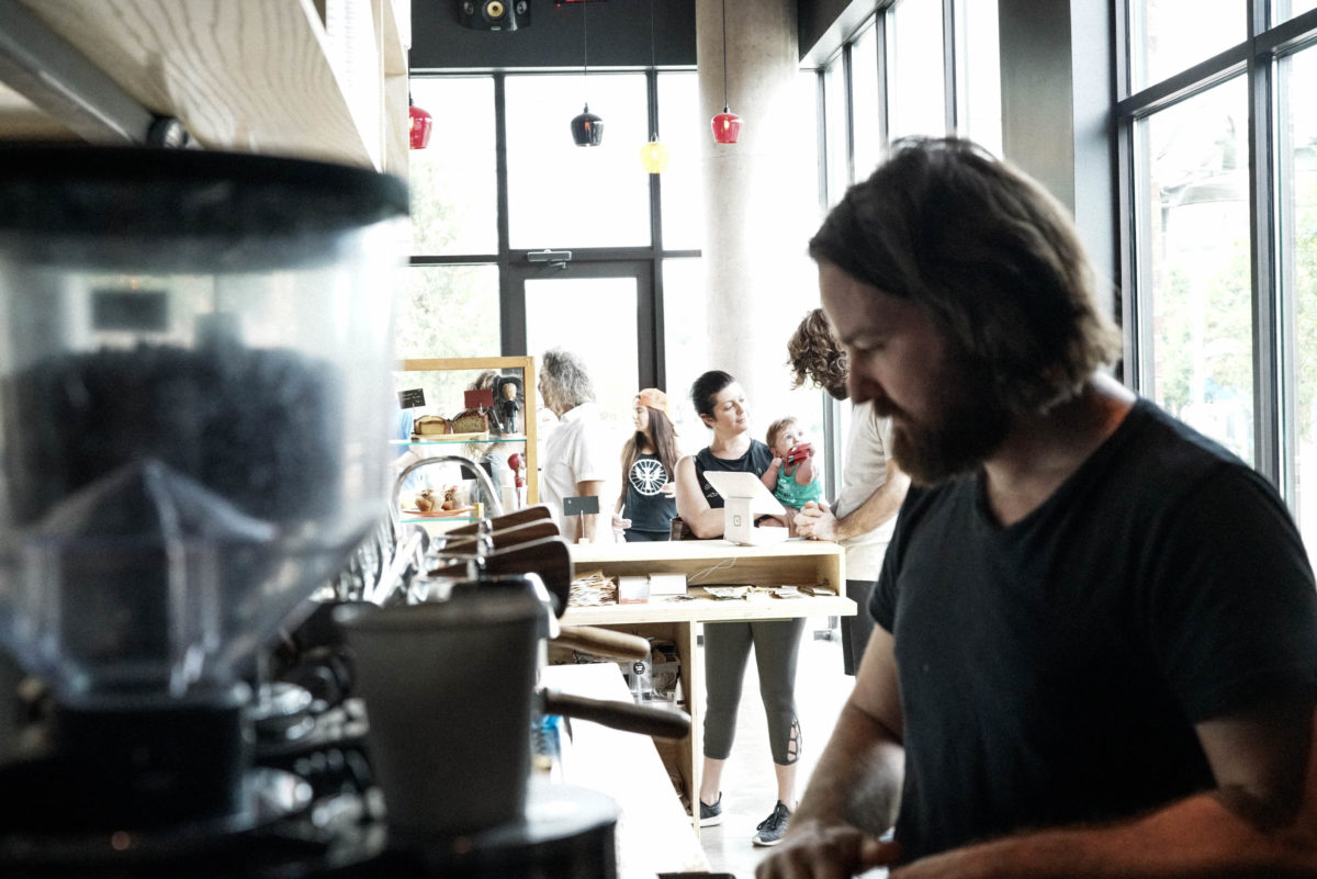 A dark foreground in which a bearded man prepares coffee and a light background in which groups of people sit in front of a bright window