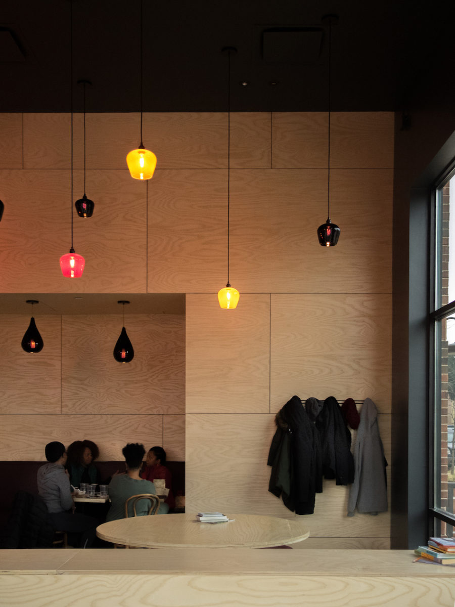 lights hang down in front of a light wood wall, beneath which sits a long row of black and gray coats to the right and a group of friends sitting to the left