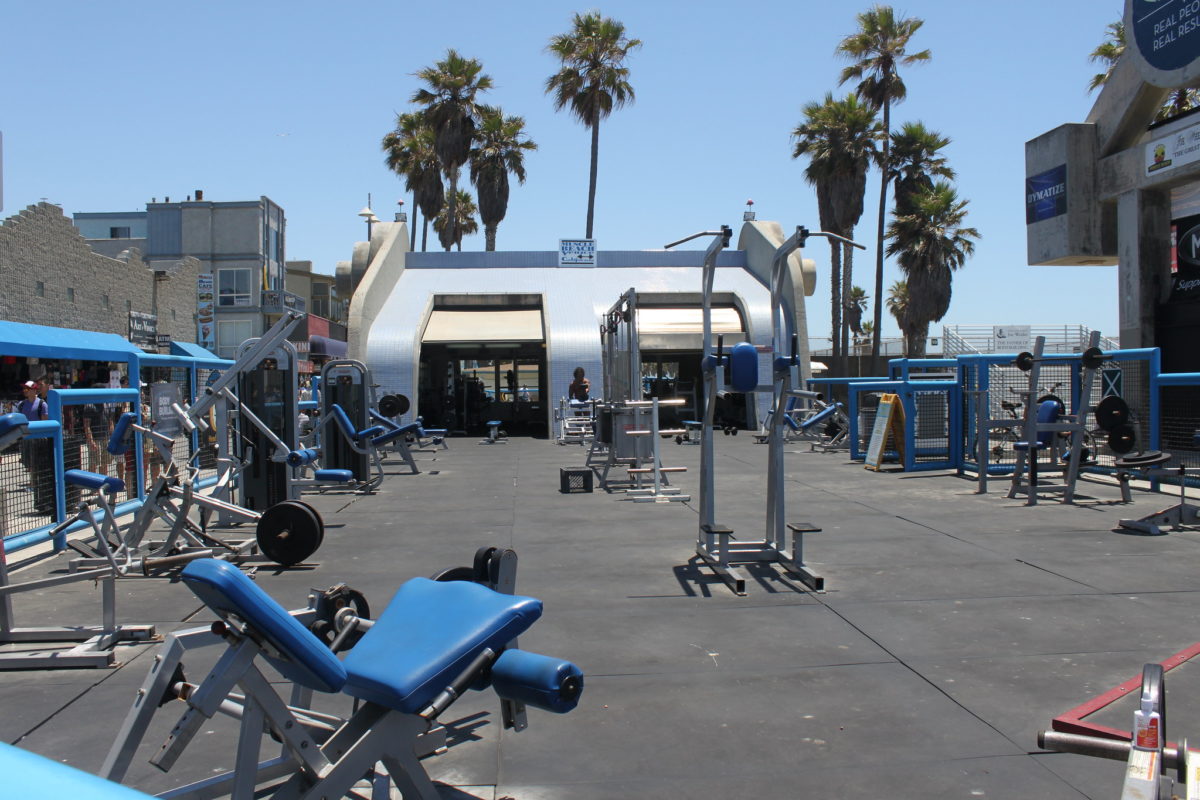 a blue trailer sits at the back of a concrete lot filled with exercise equipment with palm trees in the background