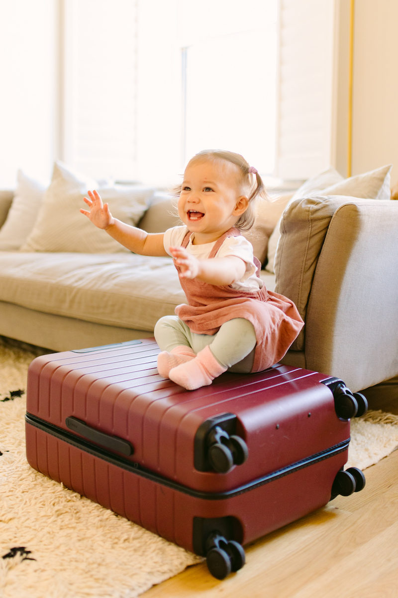 a baby sits on top of a dark red suitcase with a playful expression