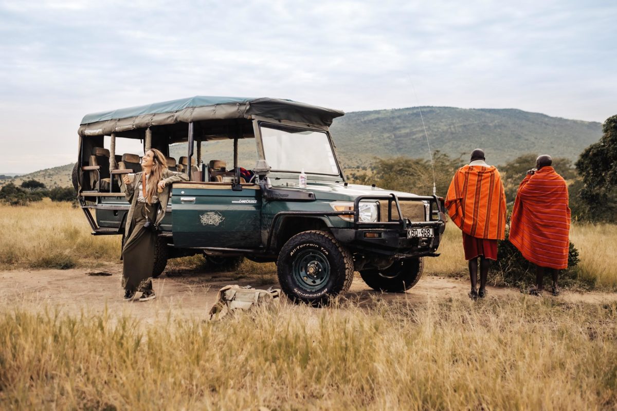 a woman stands in front of a jeep as two people in orange robes walk into a field