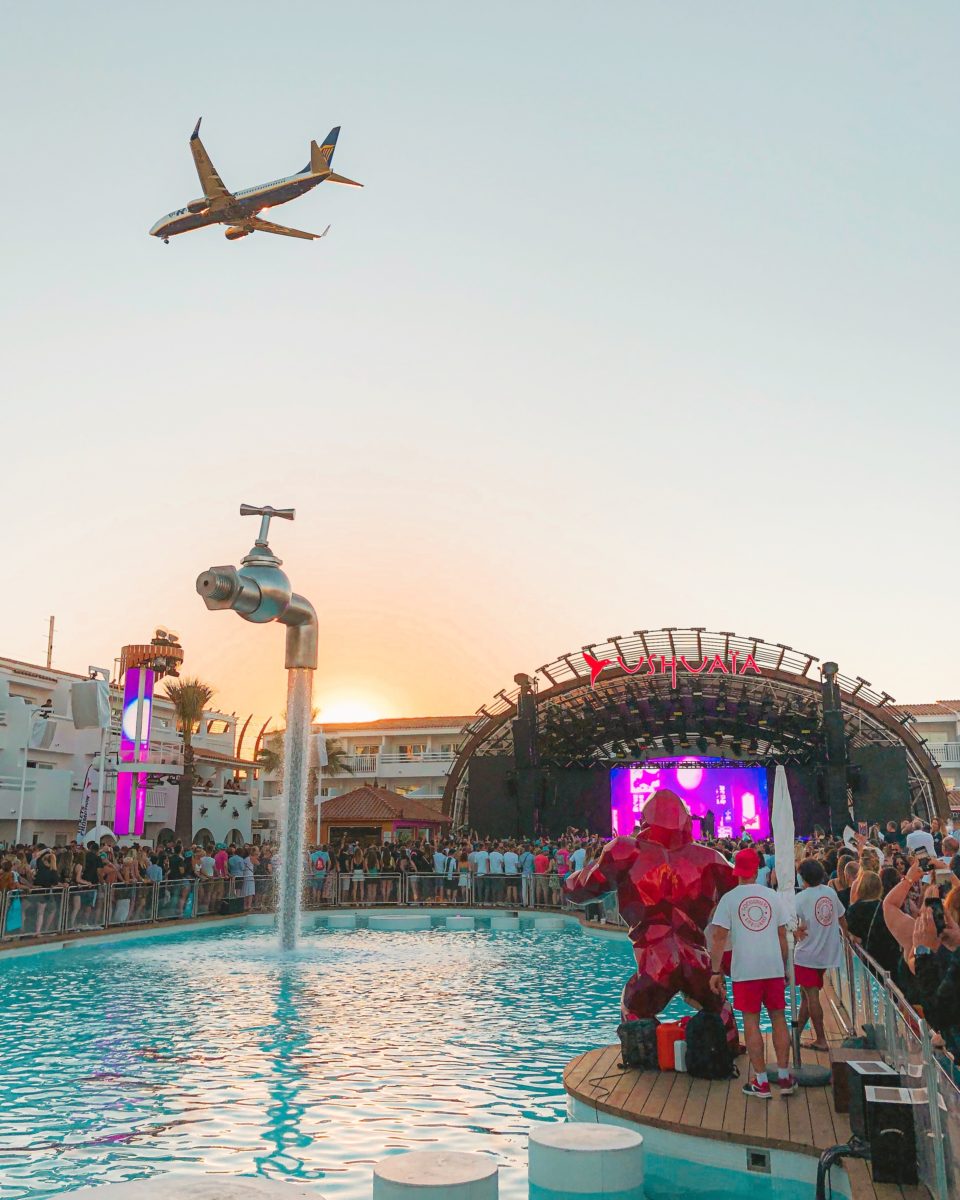 a plane soaring over an open-air club with a large pool and purple-lit stage