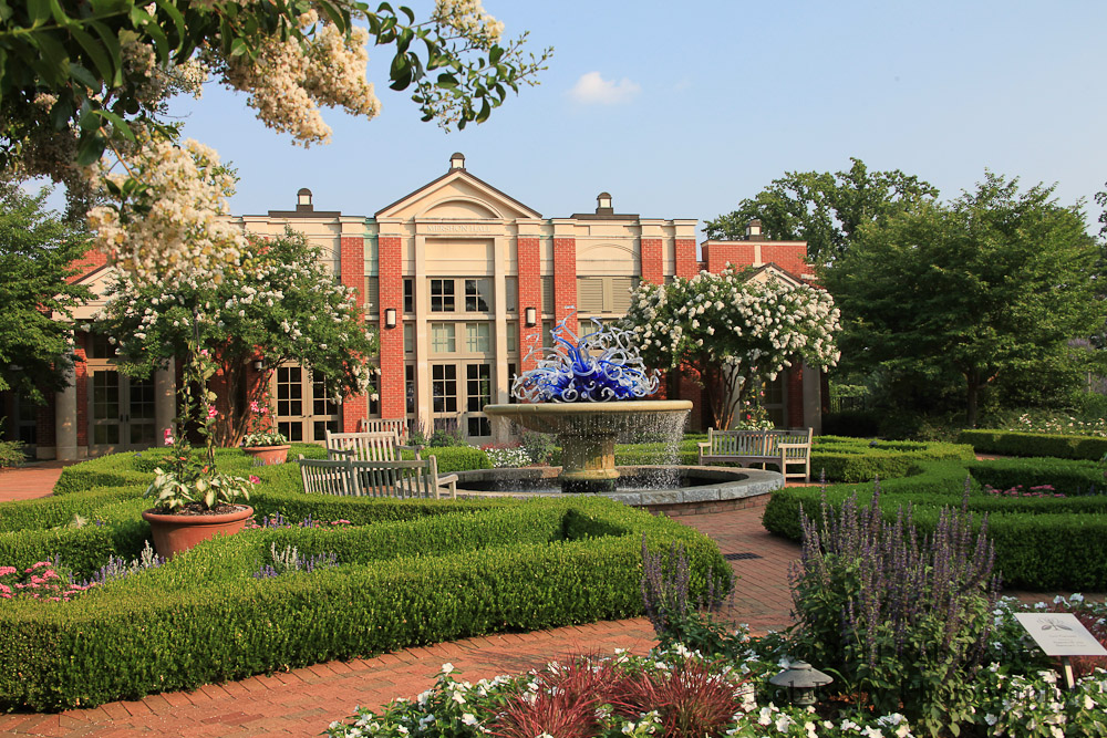 a building sitting in the middle of plaza with a fountain, hedges, a glass sculpture, and flowers