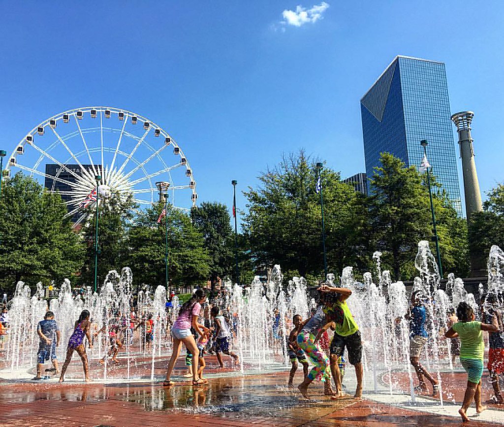 kids playing in fountains in a plaza with trees, buildings, and a ferris wheel in the background