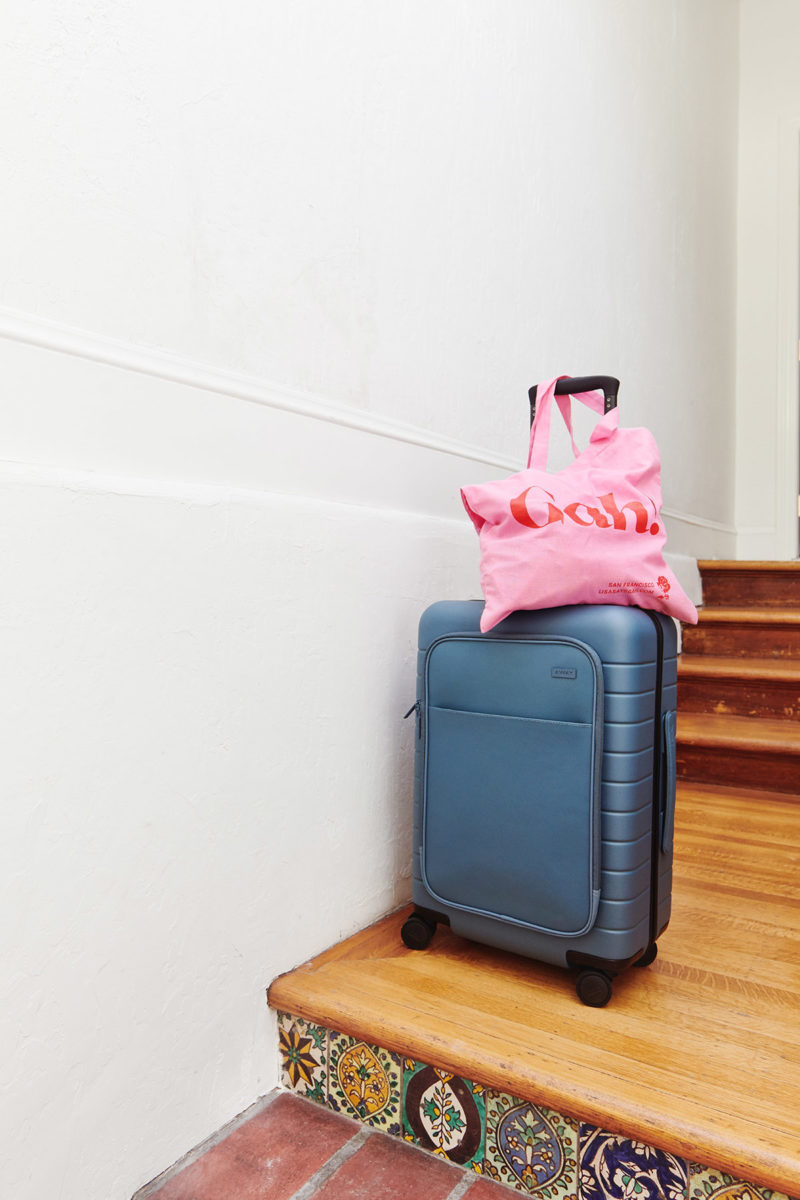 a blue Away suitcase with a pink Lisa Says Gah tote on top in a hallway