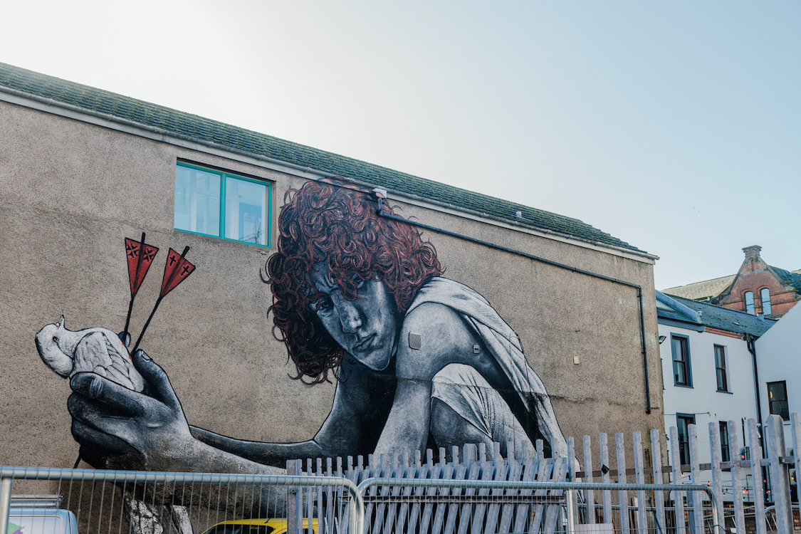 a mural of a person with curly hair crouching while holding a pigeon with two darts in its chest