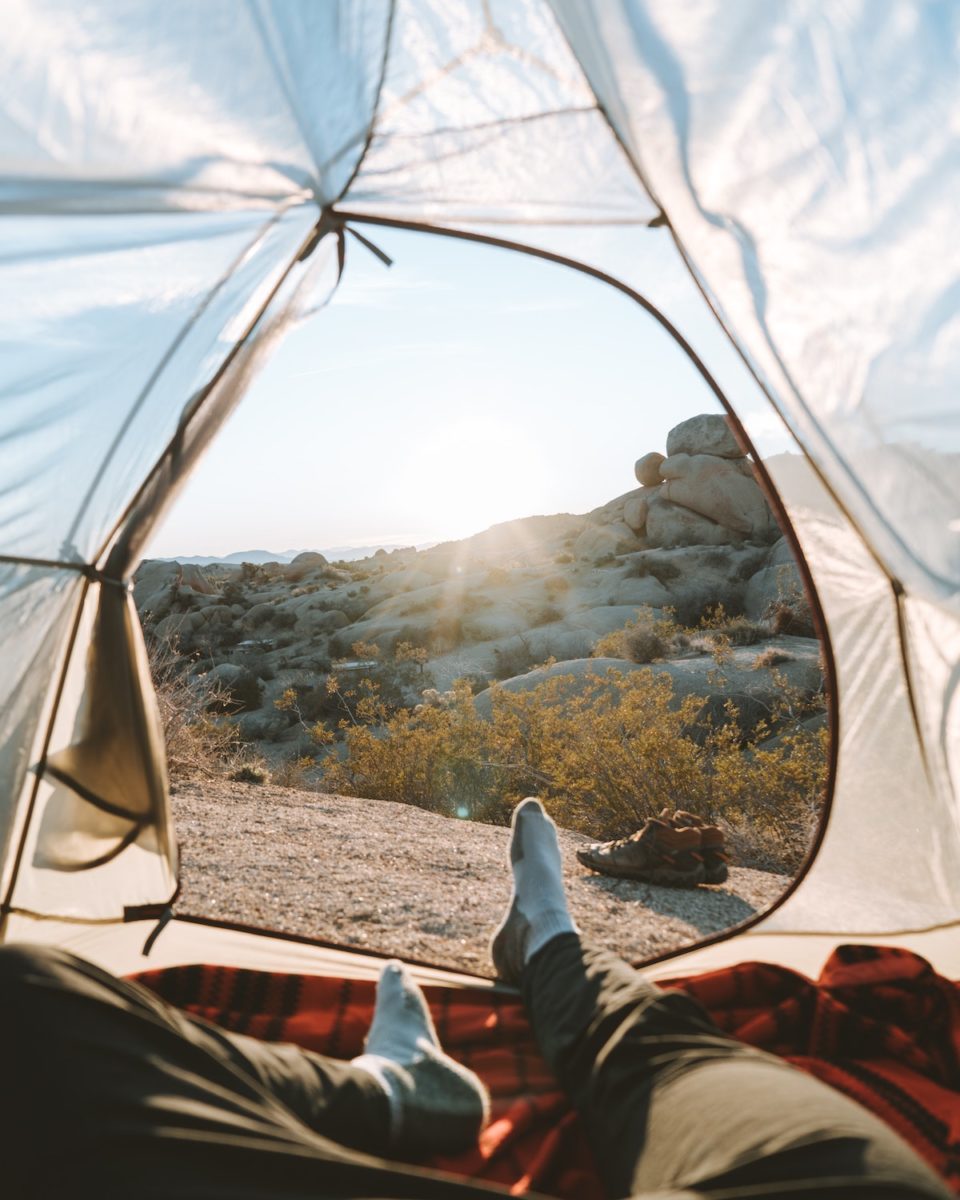 A man's legs jutting out of a white tent overlooking his campsite and greenscape