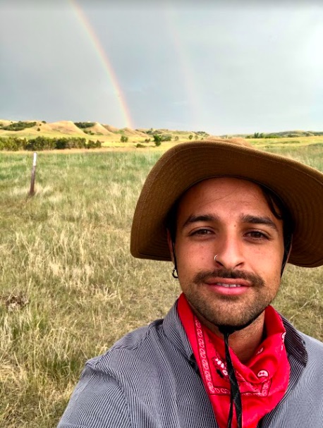 man in a hat taking a selfie in front of a pasture with a rainbow in the background